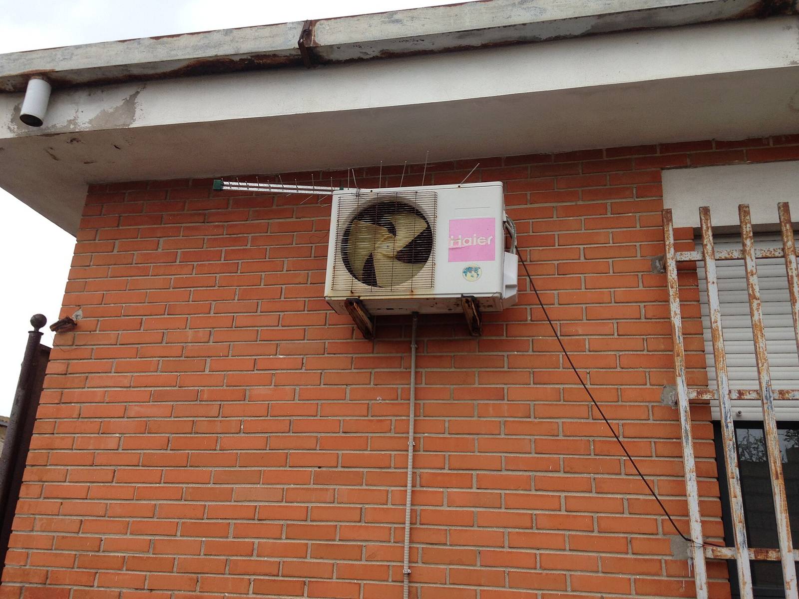 Buitenunit van een airco op een muurbeugel hoog tegen een rode bakstenen gevel, onder de dakrand en naast een raam met traliewerk