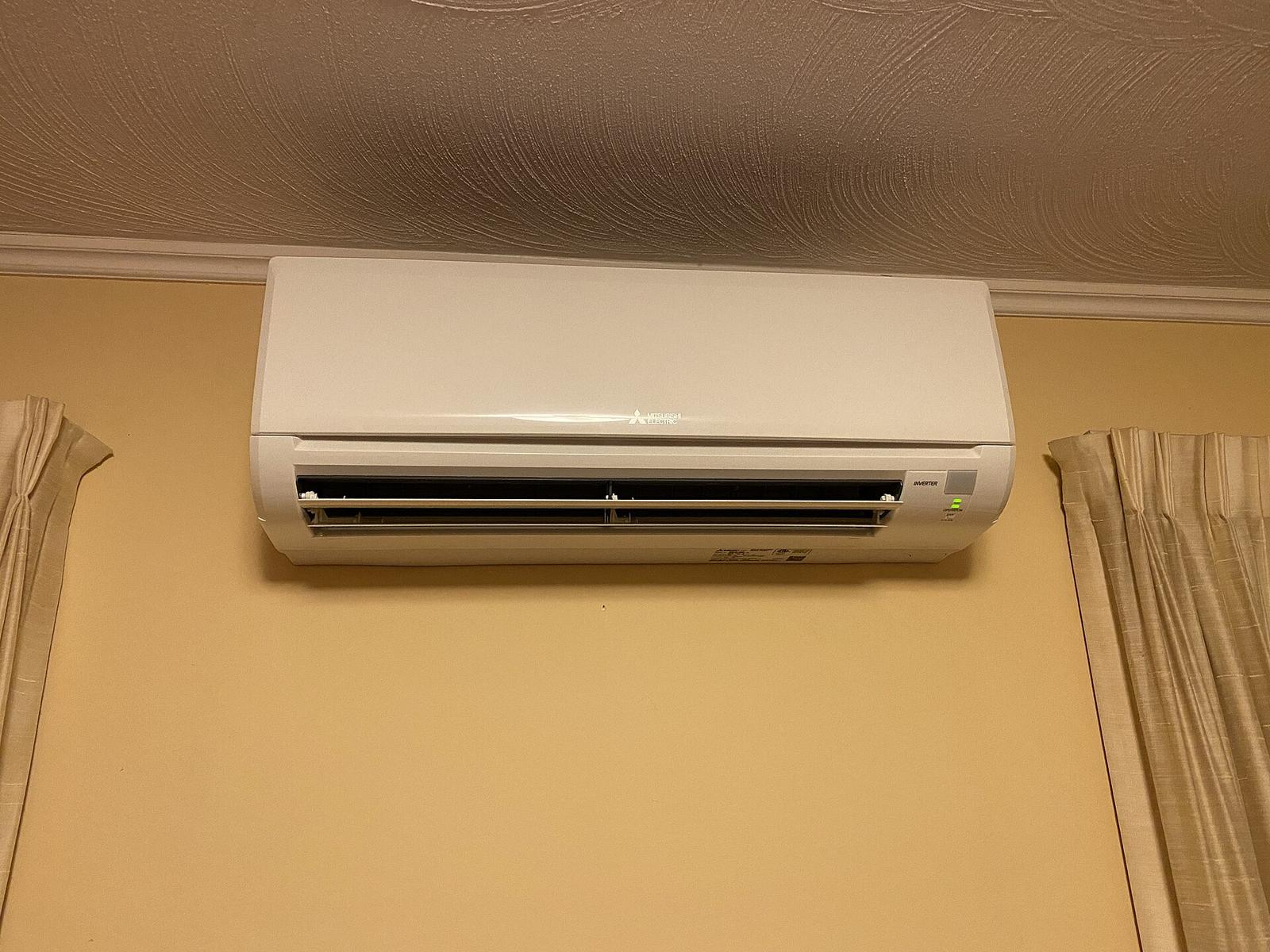 Witte binnenunit van een warmtepomp hoog tegen een okergele muur, tussen twee beige gordijnen