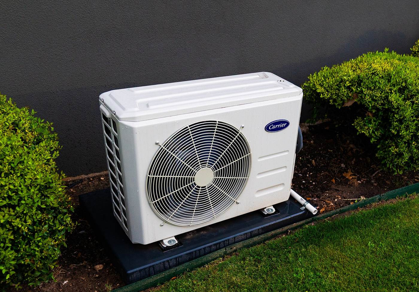 Witte buitenunit van een split-airco op een zwarte sokkel tussen twee groene struiken, tegen een donkere gevel