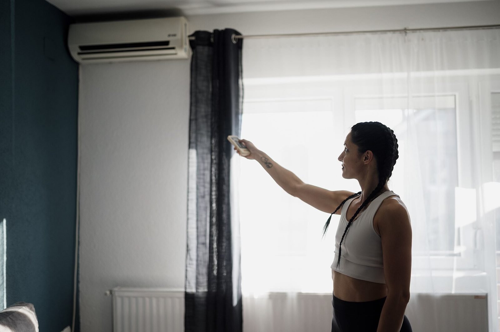Vrouw bedient met een afstandsbediening een wandairco boven een raam met gordijnen, met een radiator onder het raam
